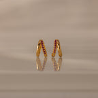 Amelia Duo Ruby Huggies—gold modern earrings with pink gemstones and intertwined hoops—stand upright on a reflective beige surface.