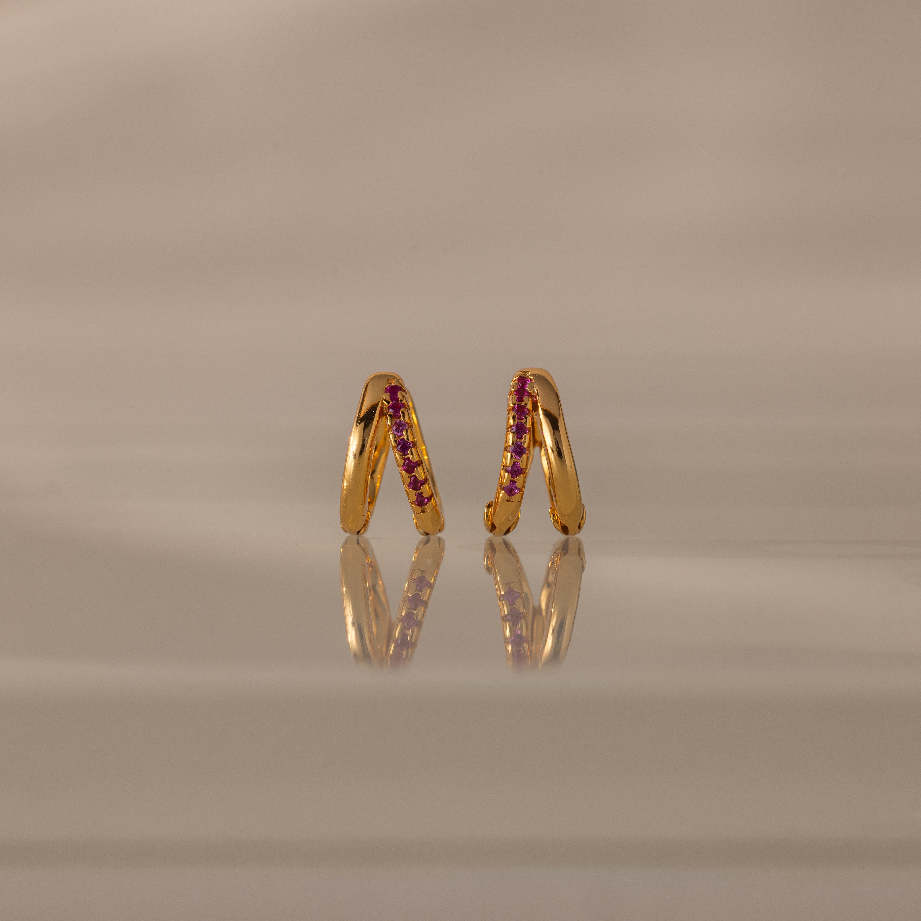 Amelia Duo Ruby Huggies—gold modern earrings with pink gemstones and intertwined hoops—stand upright on a reflective beige surface.