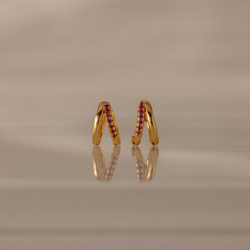 Amelia Duo Ruby Huggies—gold modern earrings with pink gemstones and intertwined hoops—stand upright on a reflective beige surface.