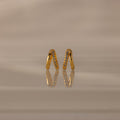 The Amelia Duo Blue Zircon Huggies, modern gold earrings featuring embedded blue zircon stones in a V shape, are showcased on a reflective beige surface.