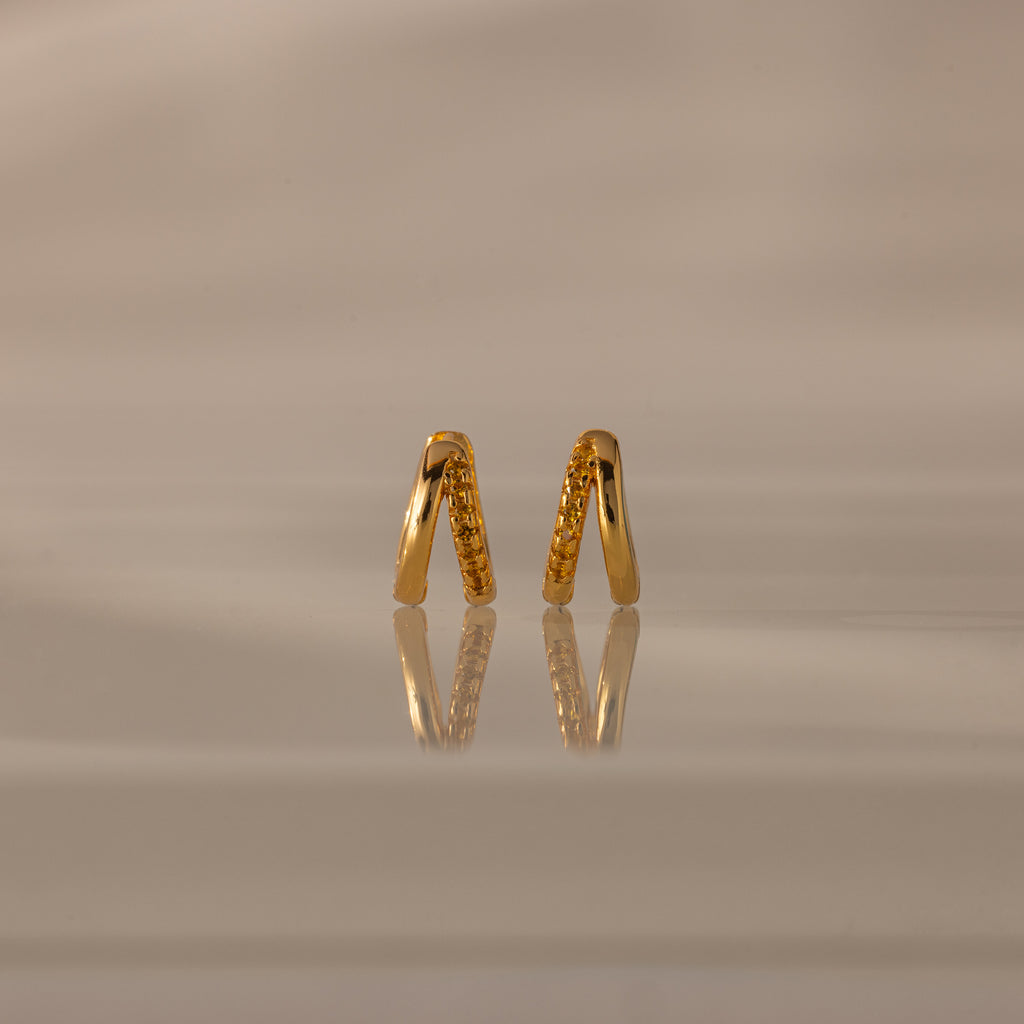 Amelia Duo Topaz Huggies, gold V-shaped modern earrings with small stones, are reflected on a glossy beige surface with a soft background.