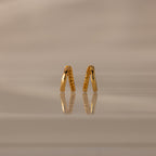 Amelia Duo Topaz Huggies, gold V-shaped modern earrings with small stones, are reflected on a glossy beige surface with a soft background.