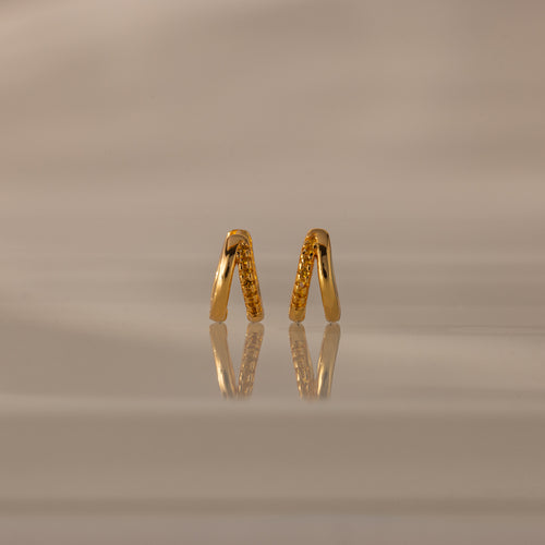 Amelia Duo Topaz Huggies, gold V-shaped modern earrings with small stones, are reflected on a glossy beige surface with a soft background.