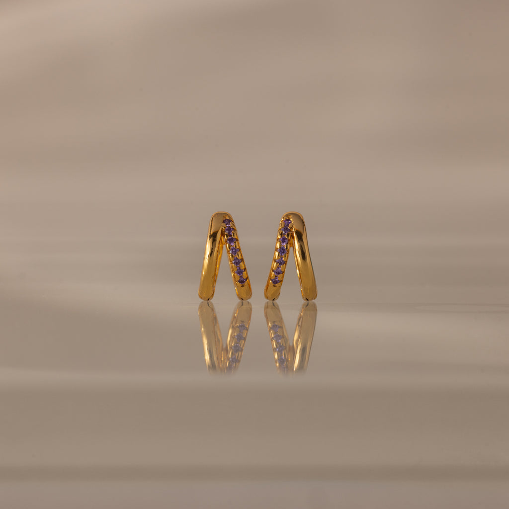 The Amelia Duo Amethyst Huggies, gold intertwined hoops with purple stones, are displayed upright on a reflective beige surface, adding a vibrant touch to your collection.