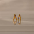 The Amelia Duo Amethyst Huggies, gold intertwined hoops with purple stones, are displayed upright on a reflective beige surface, adding a vibrant touch to your collection.
