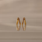 The Amelia Duo Amethyst Huggies, gold intertwined hoops with purple stones, are displayed upright on a reflective beige surface, adding a vibrant touch to your collection.