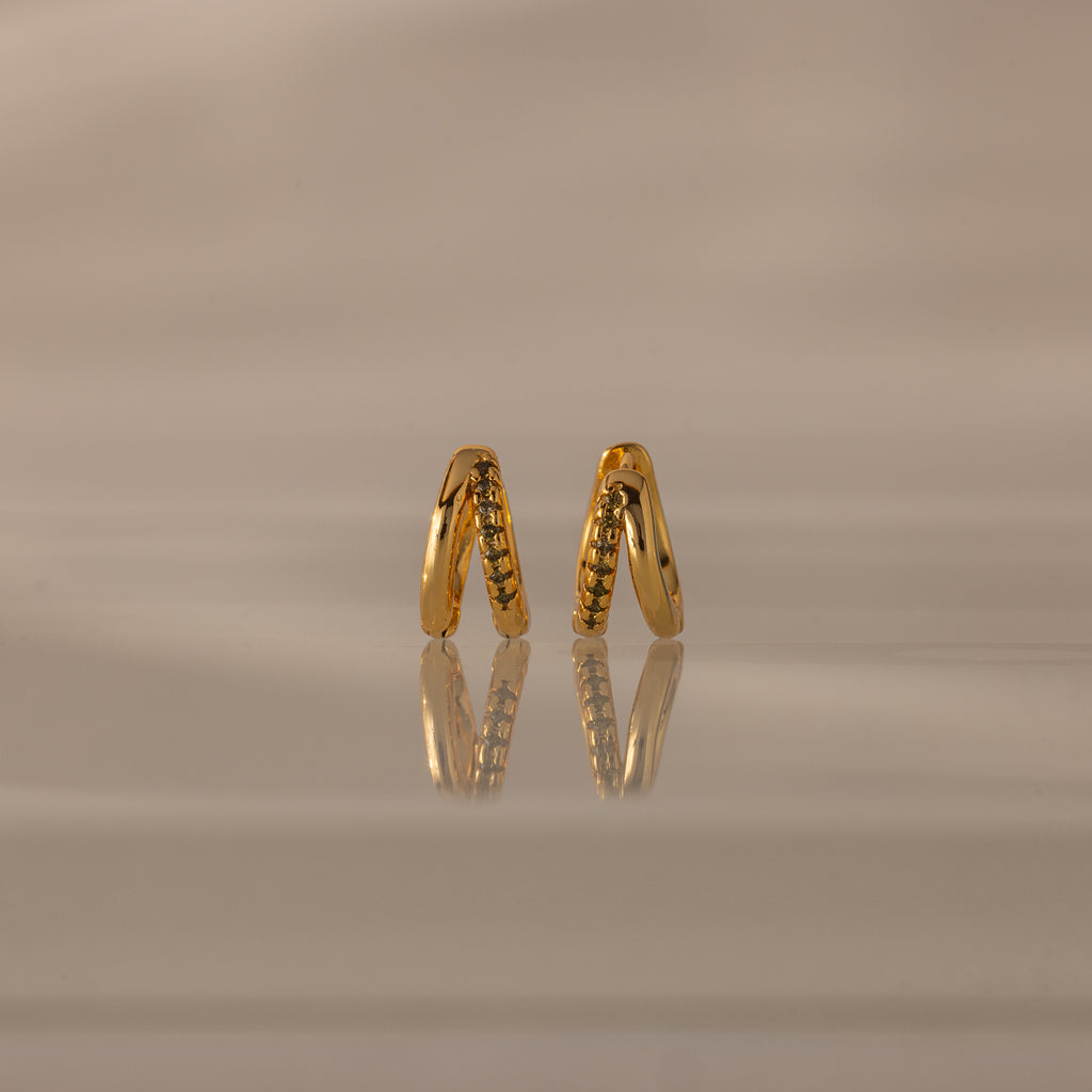 Amelia Duo Peridot Huggies—gold intertwined hoops with small peridot stones—stand upright on a reflective beige surface, offering a modern and refined take on classic earring style.