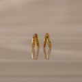 Amelia Duo Peridot Huggies—gold intertwined hoops with small peridot stones—stand upright on a reflective beige surface, offering a modern and refined take on classic earring style.