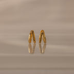 Amelia Duo Peridot Huggies—gold intertwined hoops with small peridot stones—stand upright on a reflective beige surface, offering a modern and refined take on classic earring style.
