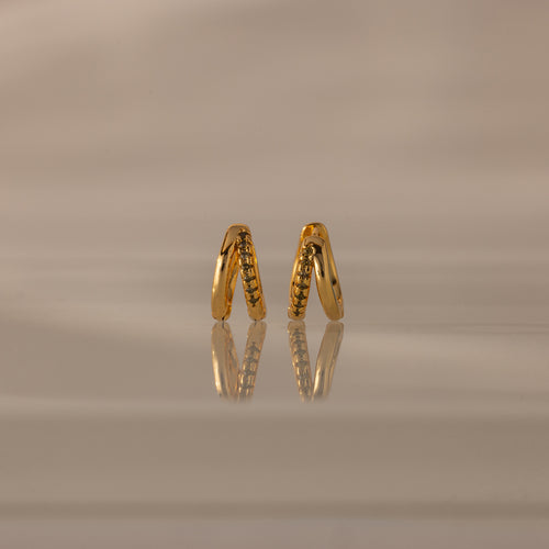 Amelia Duo Peridot Huggies—gold intertwined hoops with small peridot stones—stand upright on a reflective beige surface, offering a modern and refined take on classic earring style.