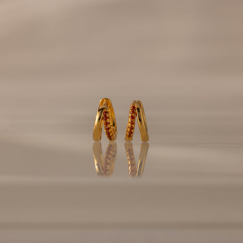 The Amelia Duo Garnet Huggies, featuring intertwined hoops and shimmering red gemstone accents, stand upright on a reflective beige surface.