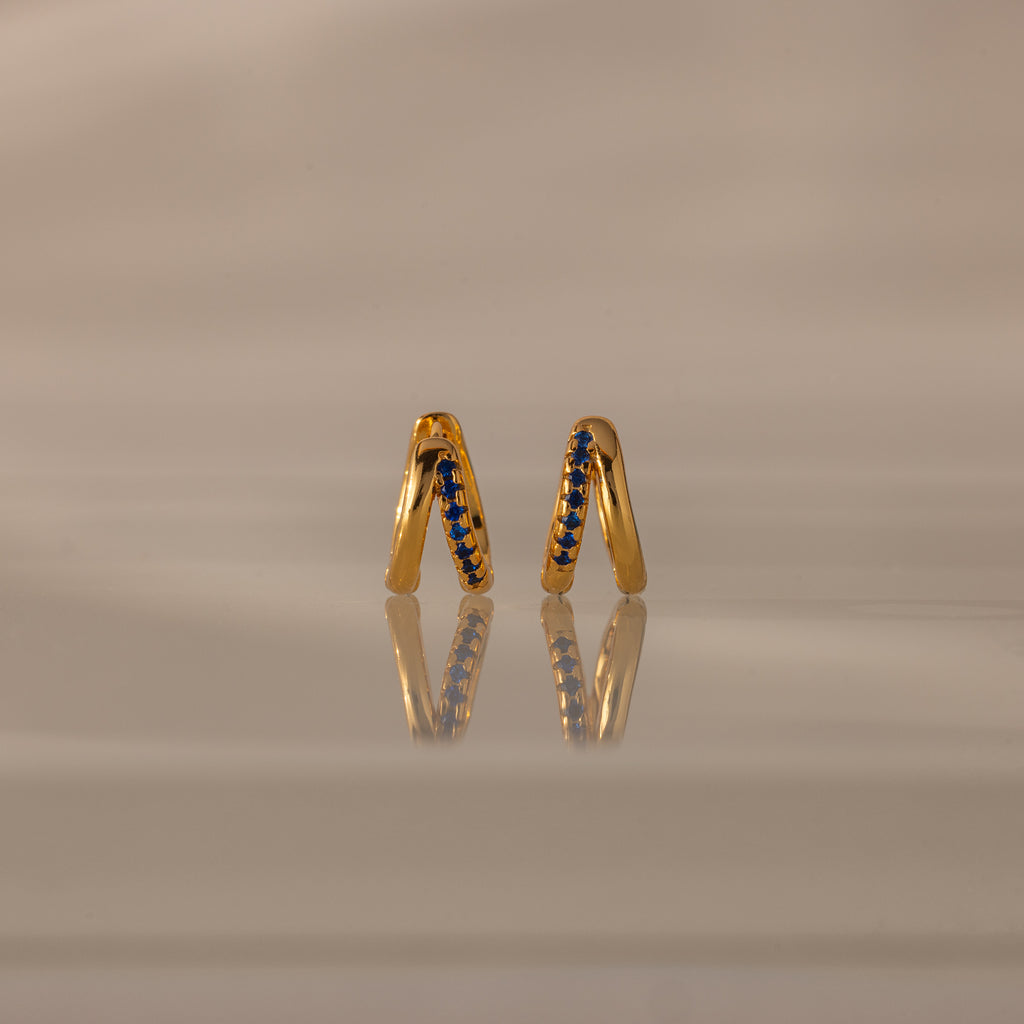 Amelia Duo Sapphire Huggies—gold intertwined hoops with blue sapphires—stand upright, reflected on a smooth, beige surface.