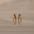 Amelia Duo Sapphire Huggies—gold intertwined hoops with blue sapphires—stand upright, reflected on a smooth, beige surface.