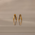 Amelia Duo Sapphire Huggies—gold intertwined hoops with blue sapphires—stand upright, reflected on a smooth, beige surface.