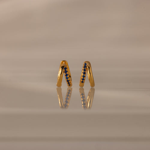 Amelia Duo Sapphire Huggies—gold intertwined hoops with blue sapphires—stand upright, reflected on a smooth, beige surface.