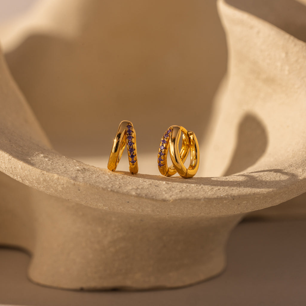 The Amelia Duo Amethyst Huggies—modern gold hoops with intertwined design and embedded amethyst stones—are displayed on a curved, textured beige surface.