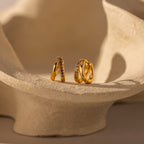 The Amelia Duo Amethyst Huggies—modern gold hoops with intertwined design and embedded amethyst stones—are displayed on a curved, textured beige surface.