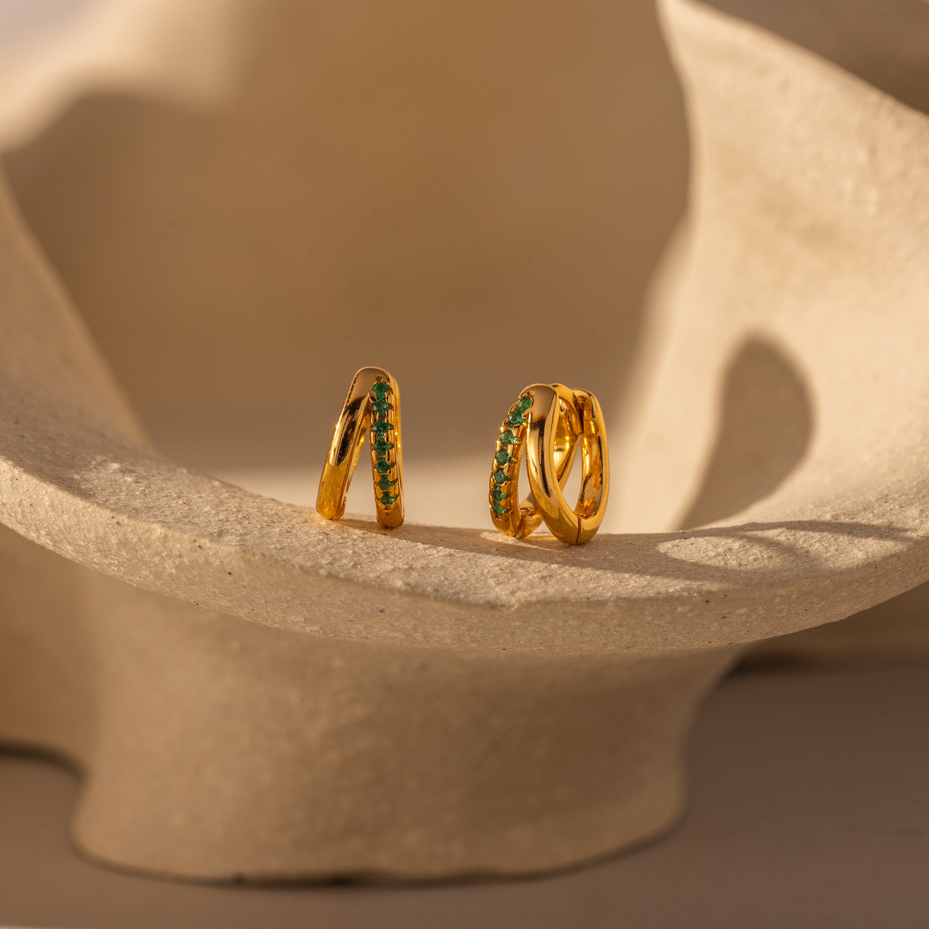 The Amelia Duo Emerald Huggies, gold intertwined hoops with green stones, are displayed on a textured, cream-colored ceramic surface.