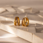 Amelia Duo Amethyst Huggies: modern gold intertwined hoop earrings adorned with small purple amethyst stones, showcased on a textured geometric beige surface.