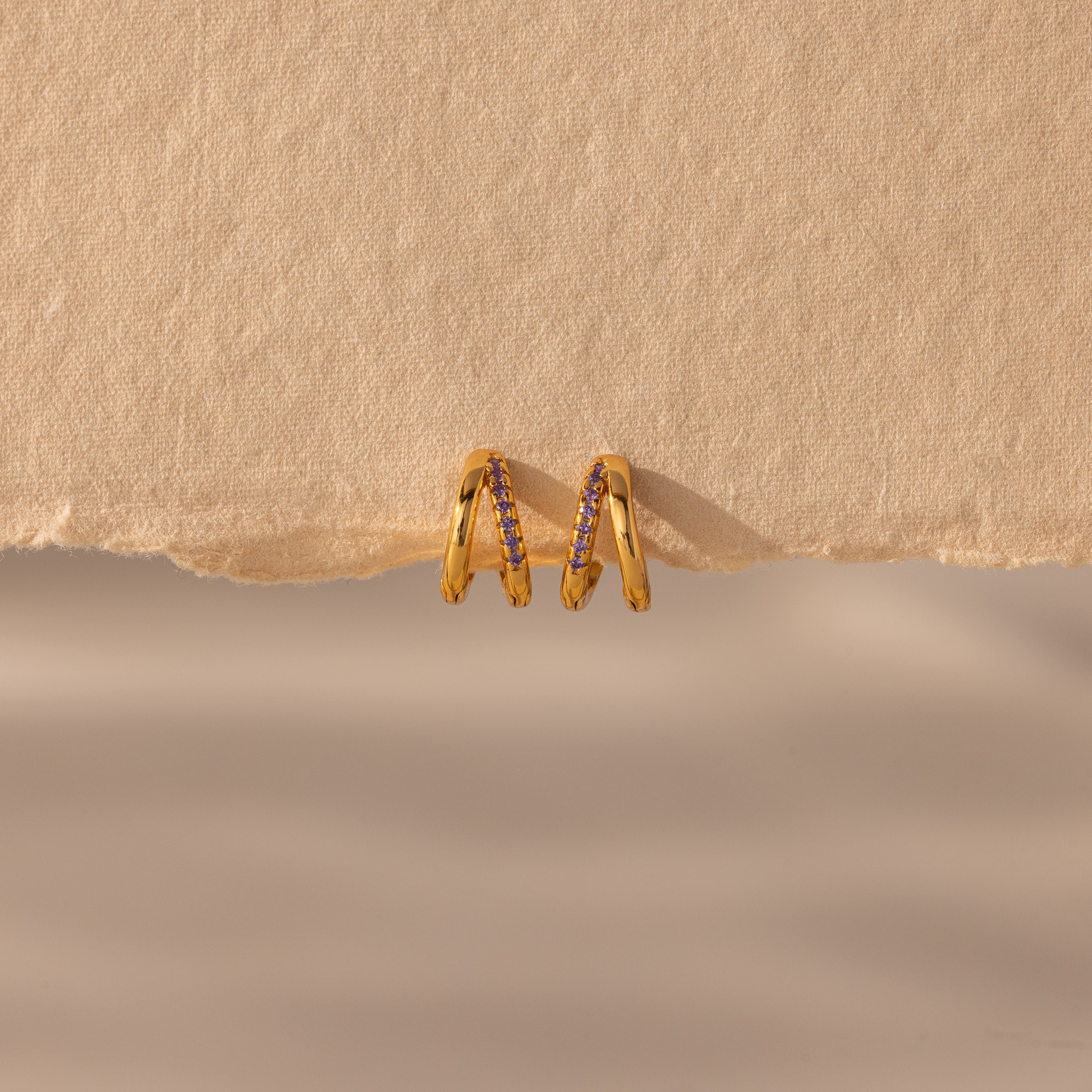 The Amelia Duo Amethyst Huggies are modern gold intertwined hoops adorned with small purple stones, showcased on a textured beige background with a torn paper edge.