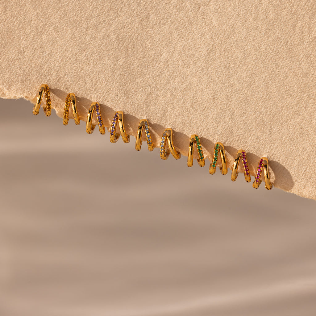 Amelia Duo Topaz Huggies—modern gold earrings with vibrant gemstones—are displayed on a torn beige paper edge against a soft background.
