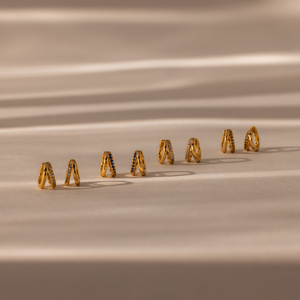 Eight pairs of earrings, including the Amelia Duo Onyx Huggies and modern intertwined hoops, are displayed on a beige surface with soft shadows.