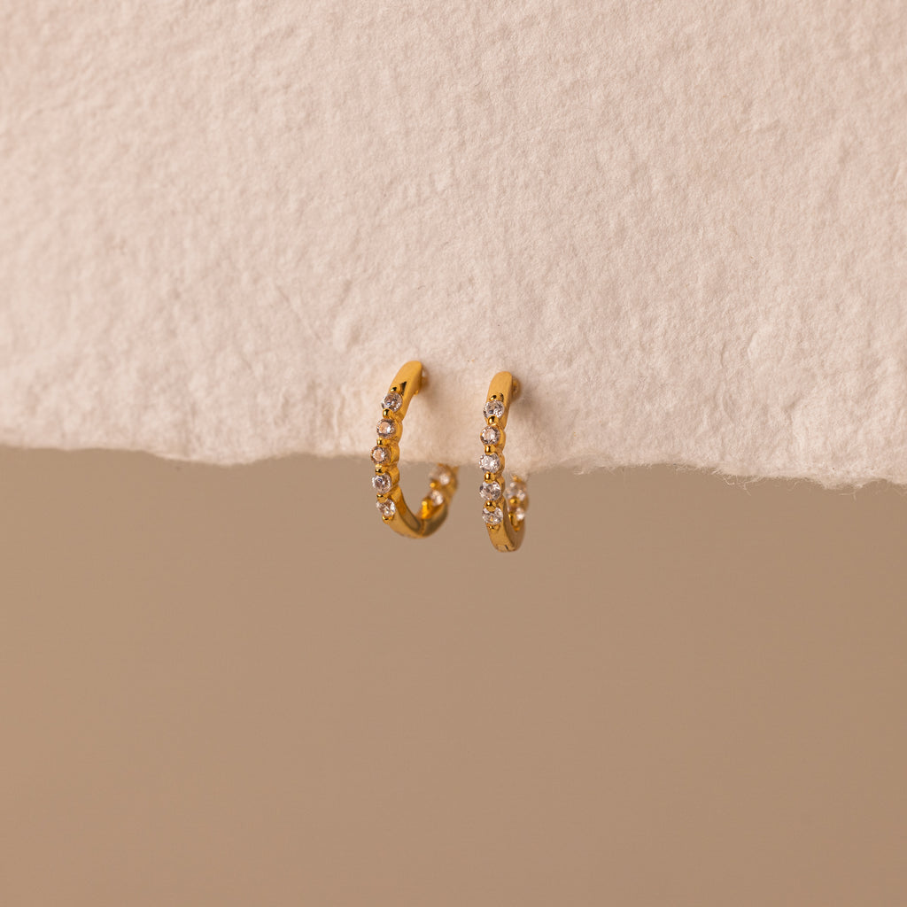 The Inside Out Diamond Huggies, gold hoop earrings adorned with sparkling diamonds, are displayed against a textured beige surface.