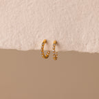 The Inside Out Diamond Huggies, gold hoop earrings adorned with sparkling diamonds, are displayed against a textured beige surface.