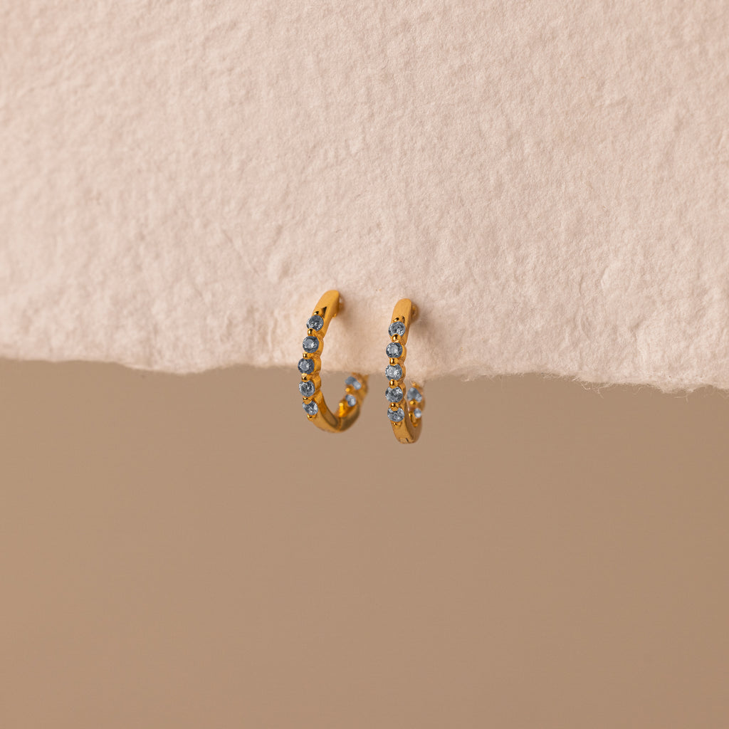 Inside Out Aquamarine Huggies: Gold hoop earrings featuring small blue aquamarine stones. Perfect for stacking or wearing solo, shown on a textured beige surface.