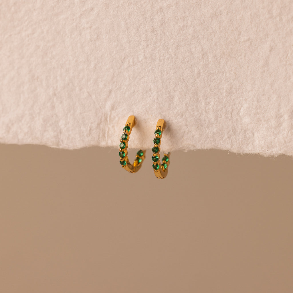 Inside Out Emerald Huggies: gold hoop earrings with small green accents, shown on a textured beige surface. Perfect as birthstone huggie earrings or for stacking to elevate your everyday jewelry style.