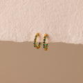 Inside Out Emerald Huggies: gold hoop earrings with small green accents, shown on a textured beige surface. Perfect as birthstone huggie earrings or for stacking to elevate your everyday jewelry style.