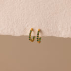 Inside Out Emerald Huggies: gold hoop earrings with small green accents, shown on a textured beige surface. Perfect as birthstone huggie earrings or for stacking to elevate your everyday jewelry style.