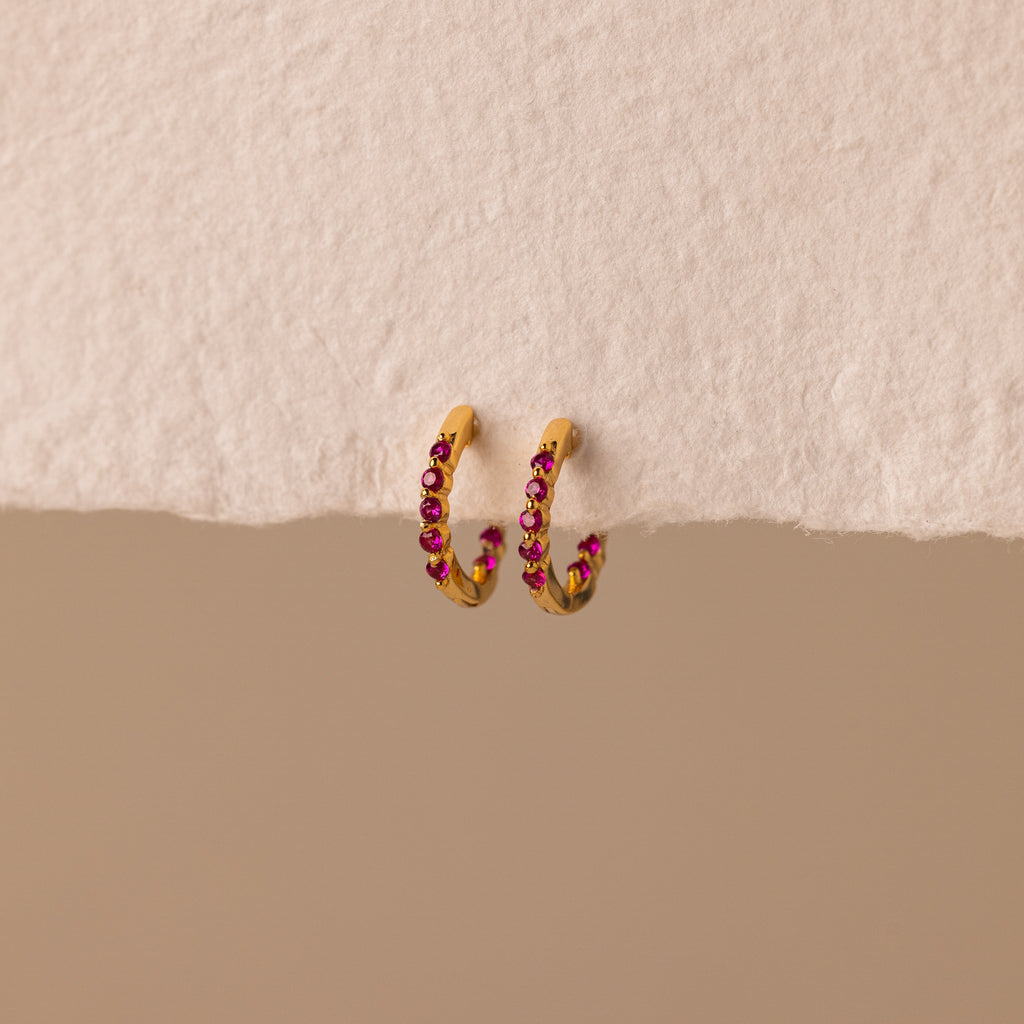 The Inside Out Ruby Huggies, featuring gold hoops with small red stones, are perfect for stacking or wearing as Birthstone Huggie Earrings and are shown on a textured off-white surface.