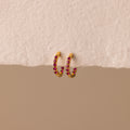 The Inside Out Ruby Huggies, featuring gold hoops with small red stones, are perfect for stacking or wearing as Birthstone Huggie Earrings and are shown on a textured off-white surface.