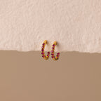 The Inside Out Ruby Huggies, featuring gold hoops with small red stones, are perfect for stacking or wearing as Birthstone Huggie Earrings and are shown on a textured off-white surface.