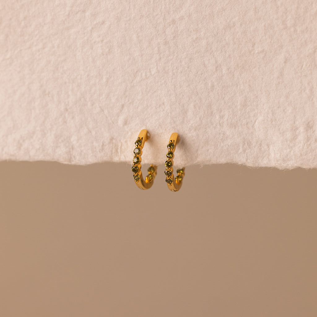 Inside Out Peridot Huggies are gold hoop stacking earrings featuring small green gemstones, perfect as birthstone huggie earrings. Shown on a textured off-white surface with a beige background.