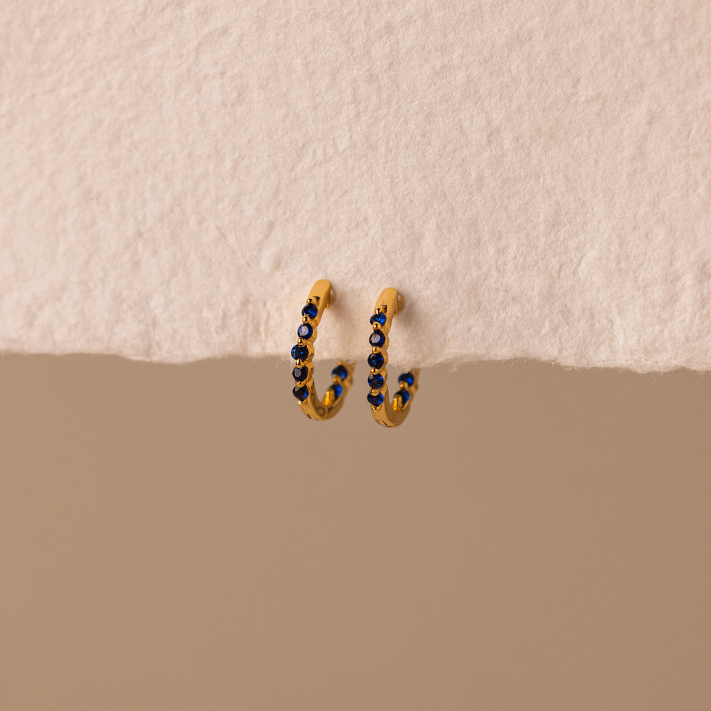 The Inside Out Sapphire Huggies feature gold hoops with small blue gemstone accents, perfect for stacking, shown on a textured off-white background.