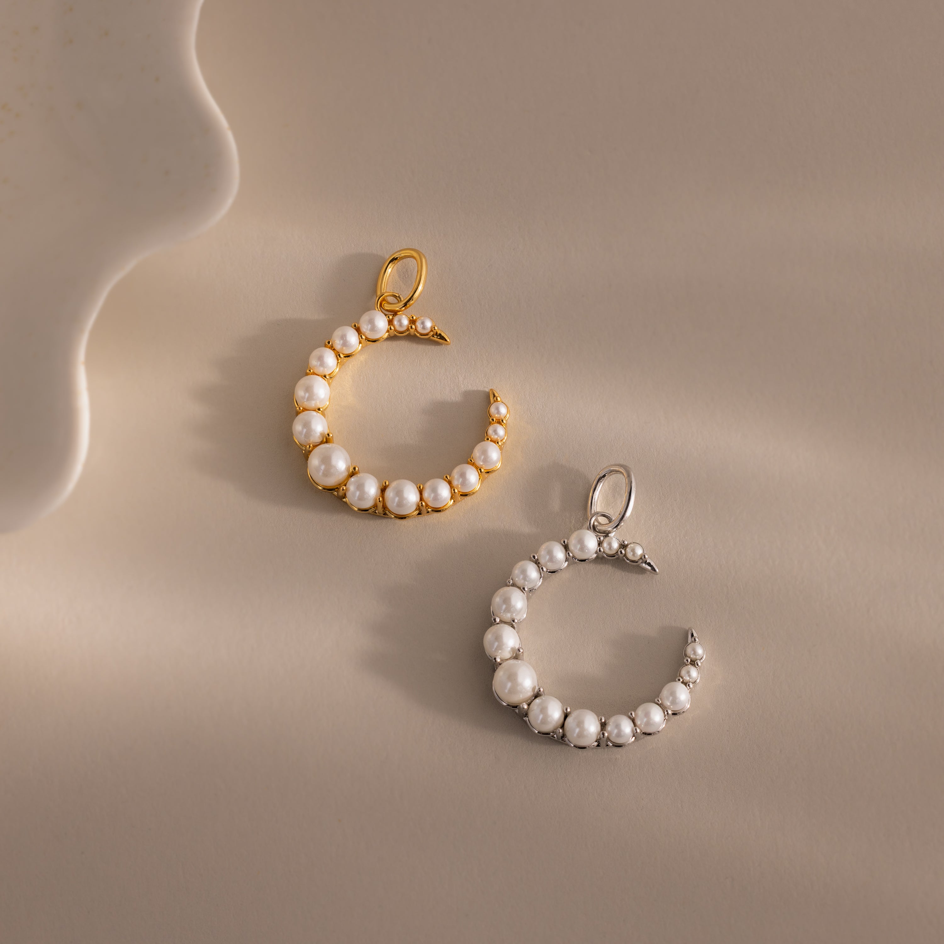 Two Pearl Moon Charm pendants—one gold, one silver—adorned with white pearls evoke celestial beauty on a beige surface.