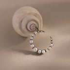 The Pearl Moon Charm in White Gold rests beside a spiral seashell on a beige surface, capturing the elegance of celestial jewelry.