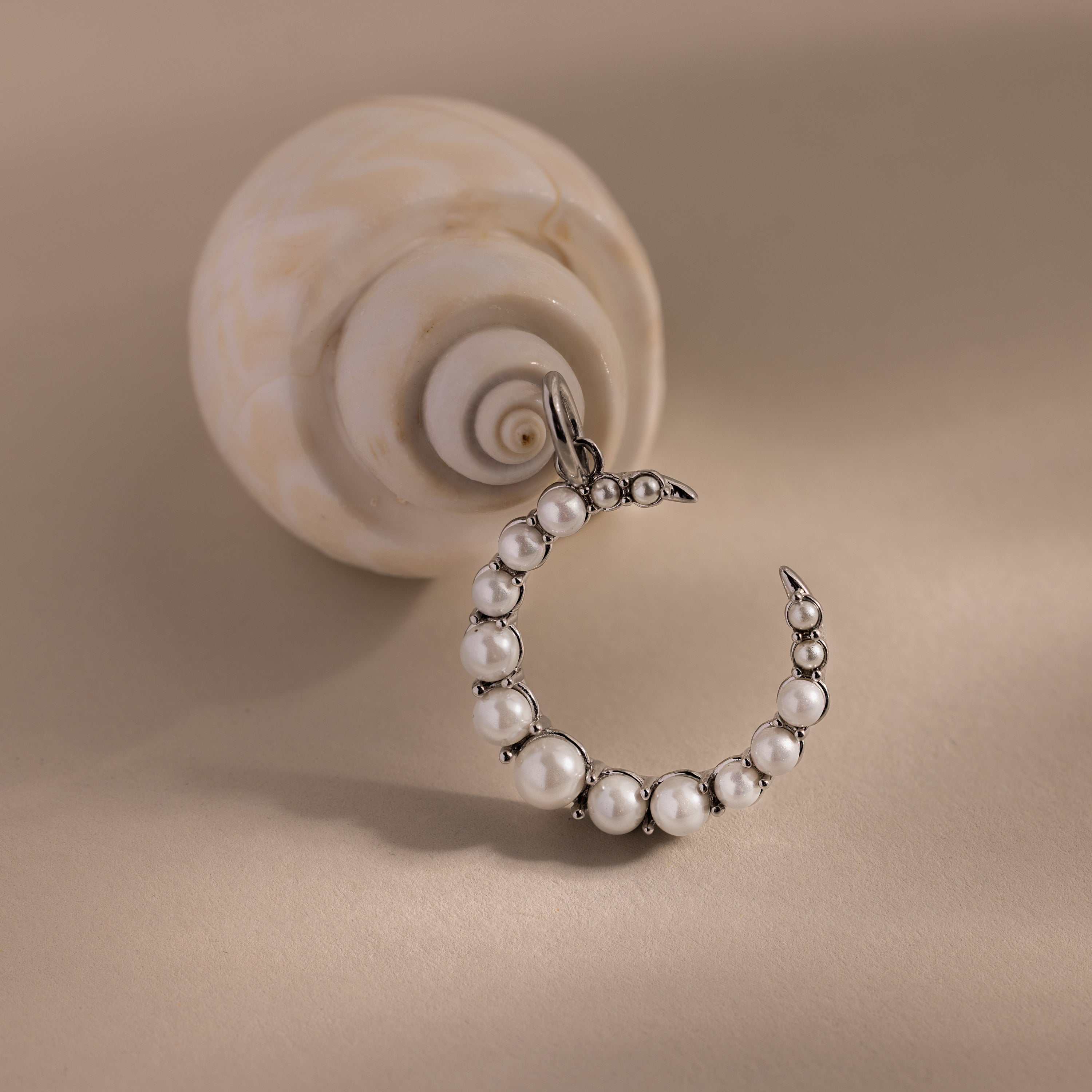 The Pearl Moon Charm in White Gold rests beside a spiral seashell on a beige surface, capturing the elegance of celestial jewelry.