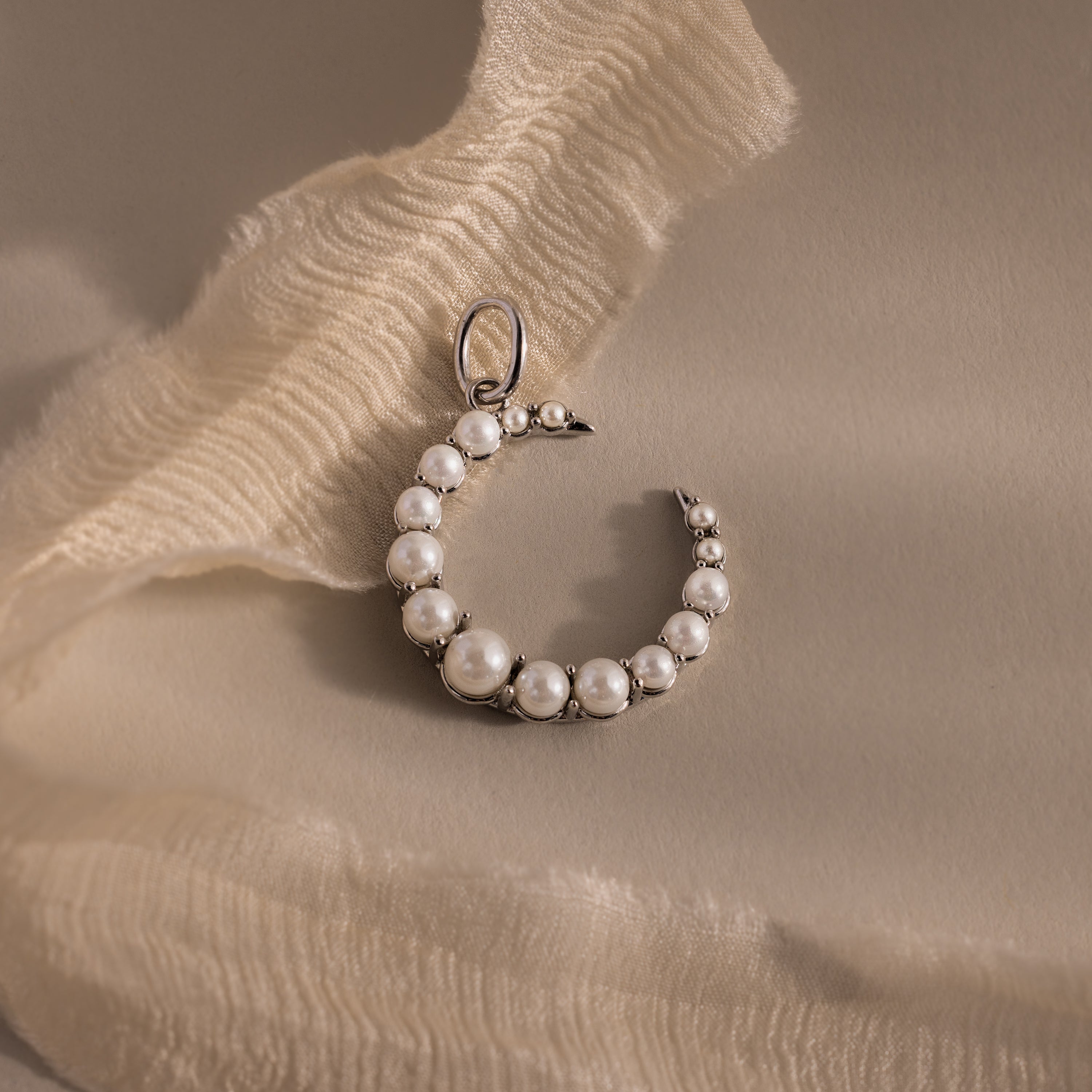 The Pearl Moon Charm, a crescent necklace accented with pearls, rests on soft cream fabric, creating an elegant celestial accessory.