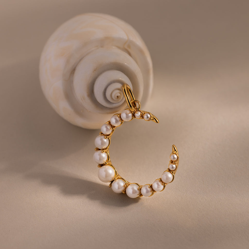 The Pearl Moon Charm, a gold crescent moon pendant with pearls, rests beside a spiral seashell on a beige surface—an elegant accessory for celestial jewelry lovers.