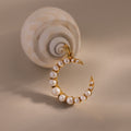 The Pearl Moon Charm, a gold crescent moon pendant with pearls, rests beside a spiral seashell on a beige surface—an elegant accessory for celestial jewelry lovers.