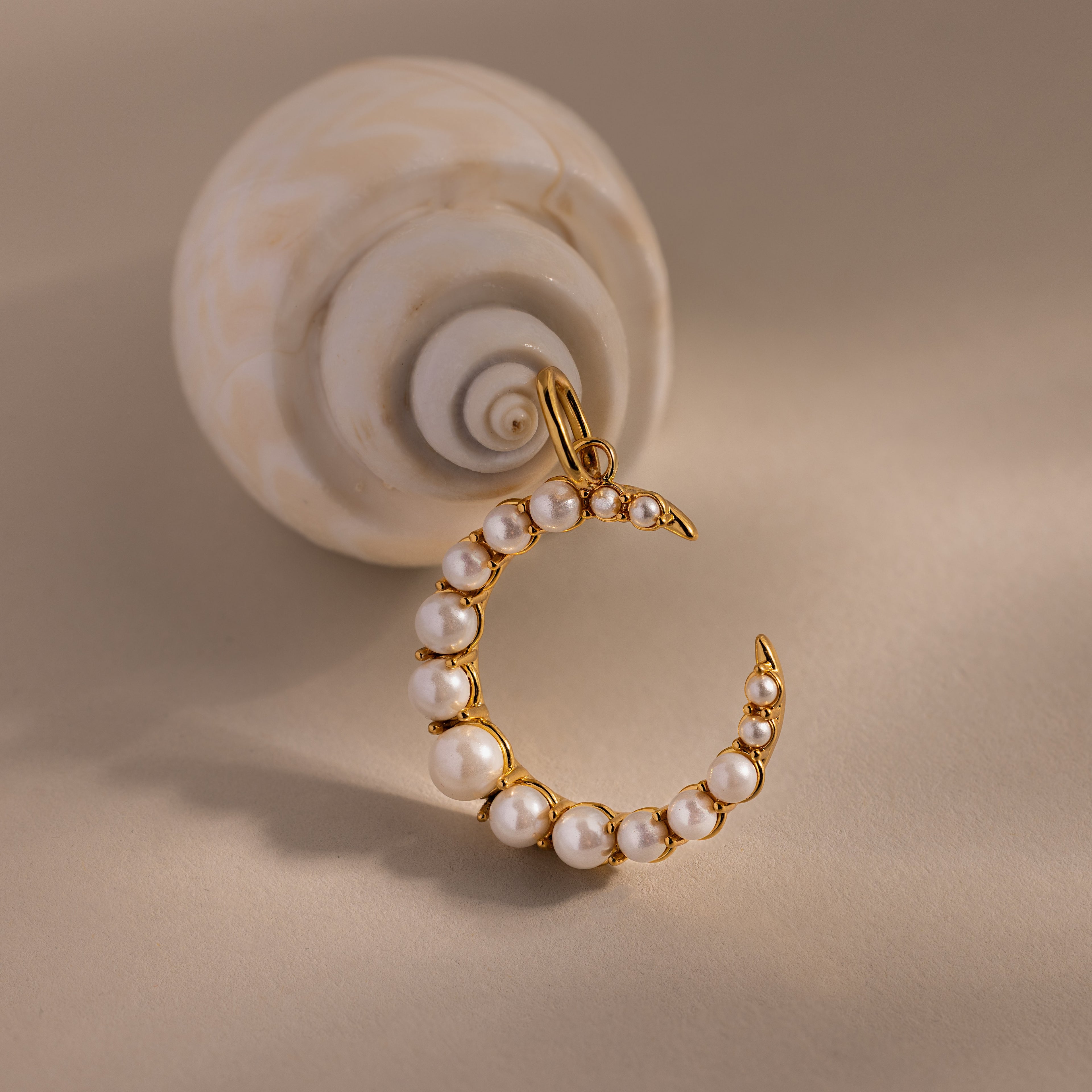 The Pearl Moon Charm, a gold crescent moon pendant with pearls, rests beside a spiral seashell on a beige surface—an elegant accessory for celestial jewelry lovers.