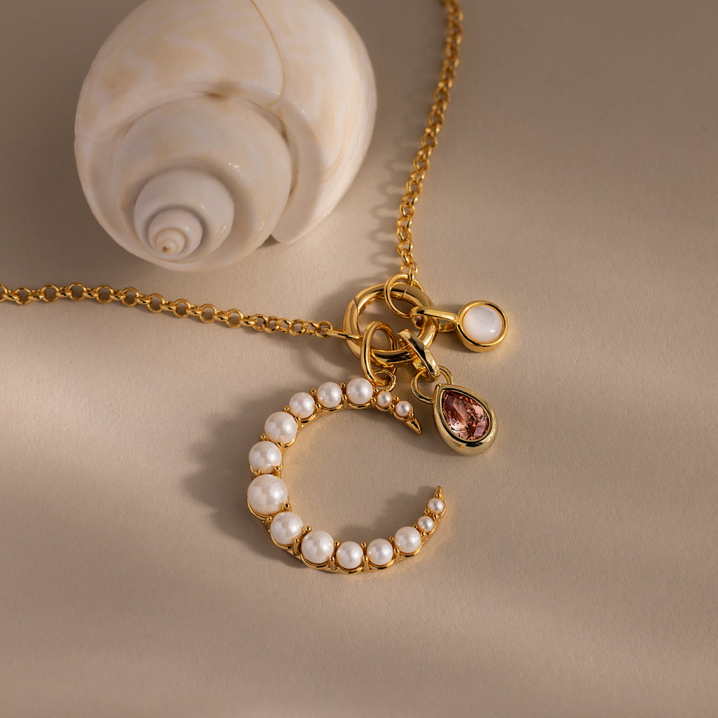 The Pearl Moon Charm in 18K Gold, adorned with delicate accents and shown beside a spiral seashell on a beige surface, is perfect for lovers of celestial-inspired jewelry.