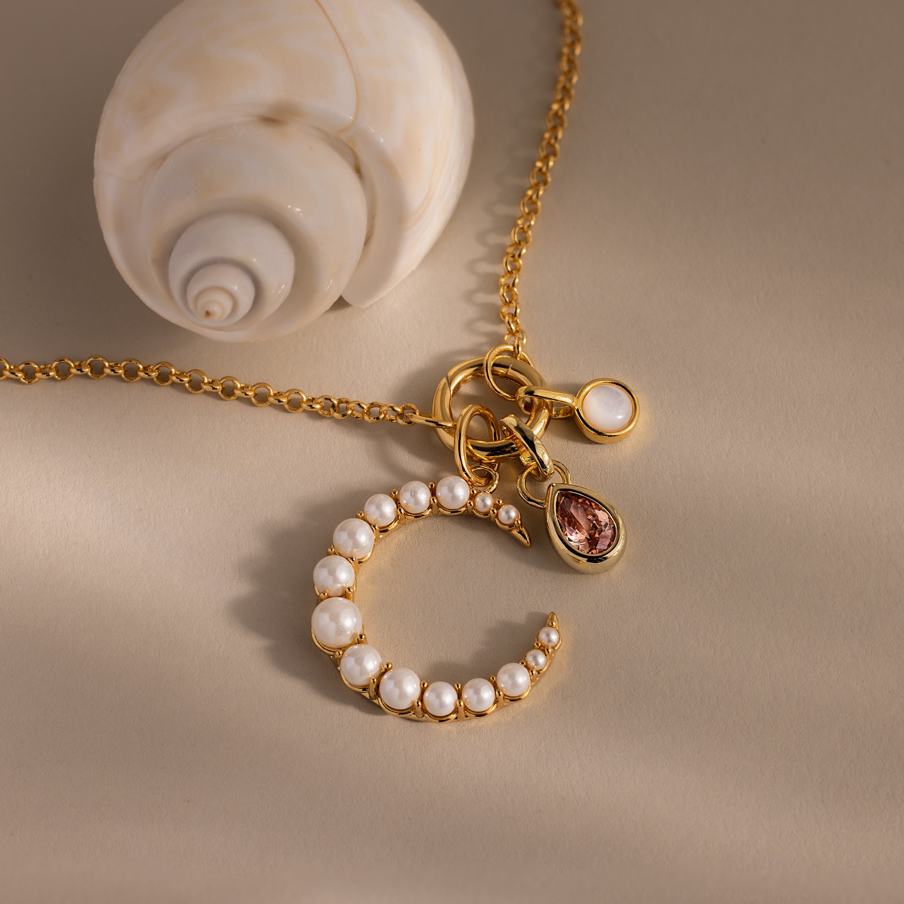 The Pearl Moon Charm in 18K Gold, adorned with delicate accents and shown beside a spiral seashell on a beige surface, is perfect for lovers of celestial-inspired jewelry.