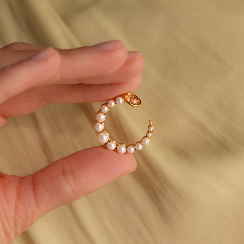 A hand displays the Pearl Moon Charm, a gold crescent-shaped ear cuff accented with delicate pearls, exuding celestial allure against a beige background.
