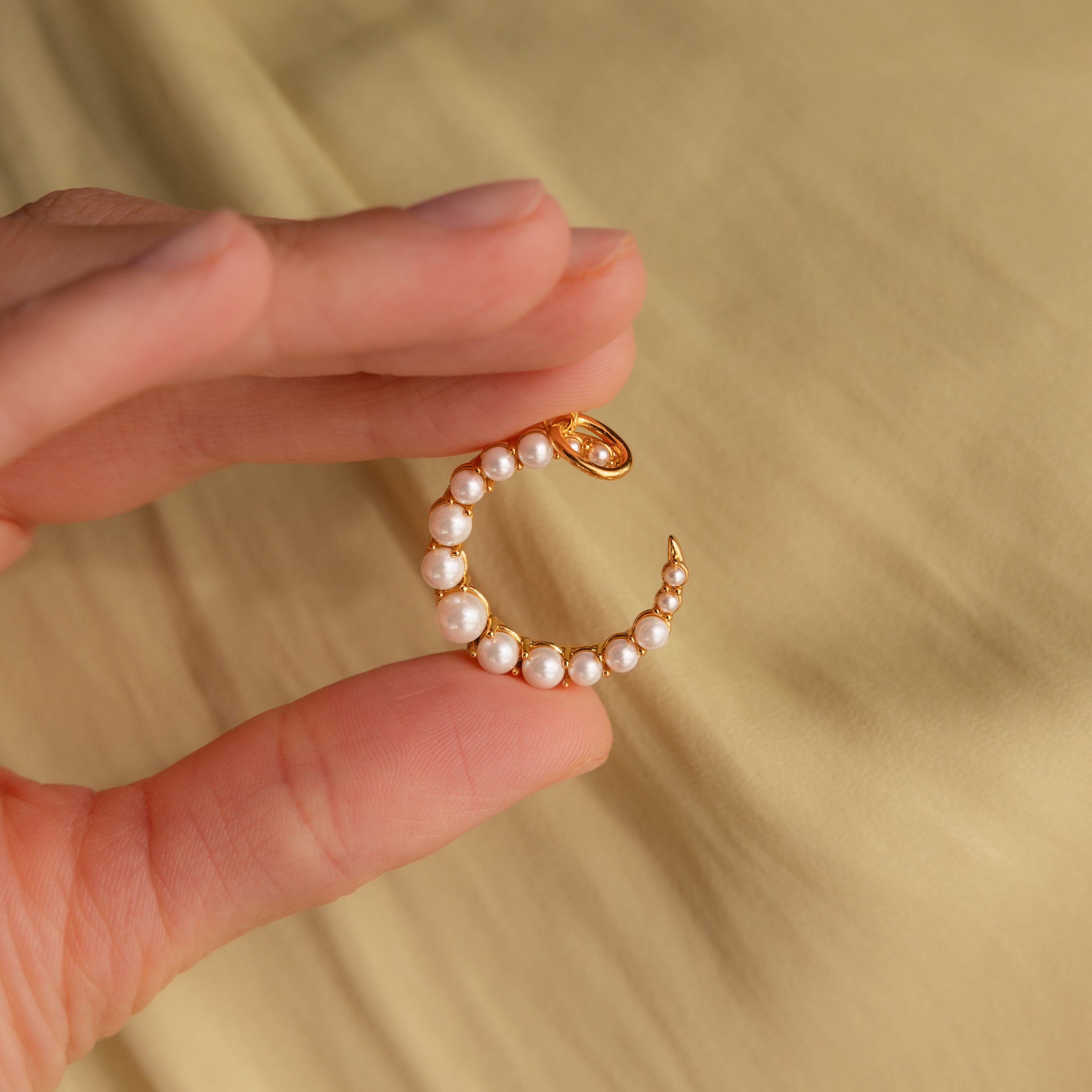 A hand displays the Pearl Moon Charm, a gold crescent-shaped ear cuff accented with delicate pearls, exuding celestial allure against a beige background.