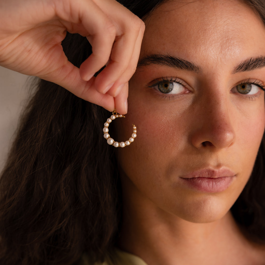A woman holds a Pearl Moon Charm gold hoop earring with pearls near her face, gazing at the camera—an elegant tribute to celestial-inspired jewelry.