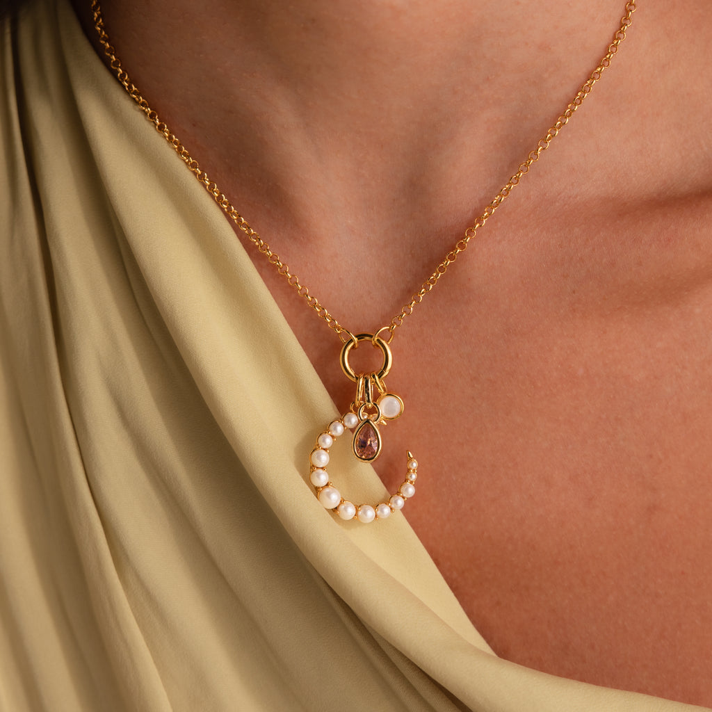 The Pearl Moon Charm gold necklace features a delicate crescent shape adorned with pearls, displayed on cream-draped fabric for a celestial, radiant look.